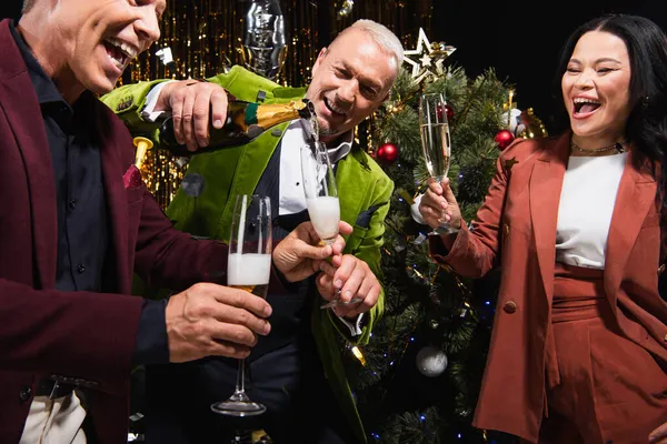 Mature man pouring champagne near excited interracial friends and christmas tree during new year party on black background — Stock Photo