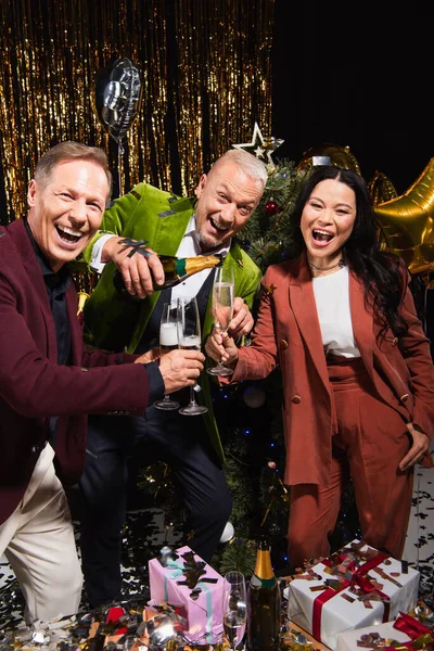 Positive interracial friends pouring champagne near christmas tree and presents on black background — Stock Photo