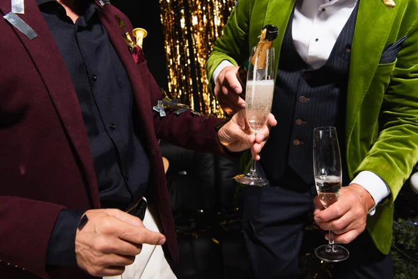 Vista recortada del hombre sosteniendo champán cerca de amigo con botella y confeti durante la fiesta sobre fondo negro - foto de stock