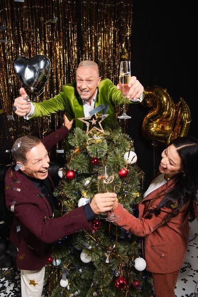 Hombre alegre sosteniendo champán y mostrando como cerca de amigos interracial y árbol de Navidad sobre fondo negro - foto de stock