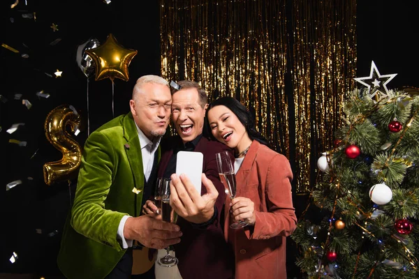 Excited multiethnic friends with champagne taking selfie on smartphone during new year party on black background — Stock Photo