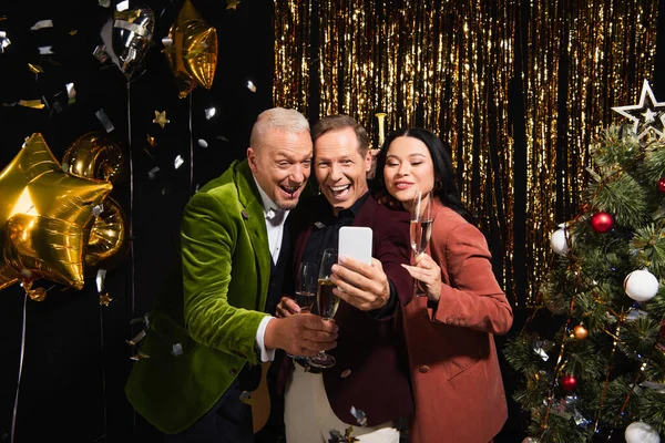 Sorrindo amigos multiétnicos com champanhe tomando selfie no smartphone perto da árvore de natal e balões no fundo preto — Fotografia de Stock