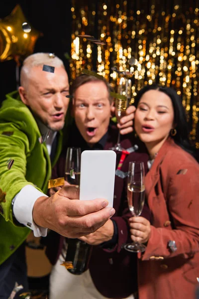 Smartphone en la mano del hombre borroso tomando selfie con amigos interracial y champán durante la fiesta de año nuevo sobre fondo negro - foto de stock