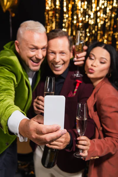 Blurred man with champagne taking selfie on smartphone with interracial friends during party on black background — Stock Photo