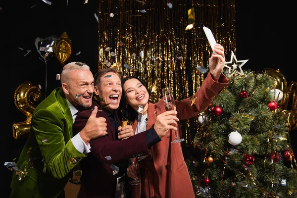 Happy interracial friends with champagne taking selfie on smartphone near christmas tree and confetti on black background — Stock Photo