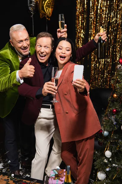 Aufgeregte interrassische Freunde machen Selfie auf Smartphone und halten Champagner in der Nähe des Weihnachtsbaums während der Party auf schwarzem Hintergrund — Stockfoto