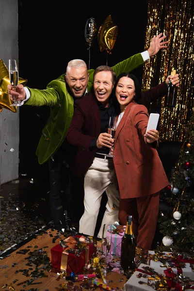 Positive interracial friends with smartphone holding champagne while celebrating new year on black background — Stock Photo