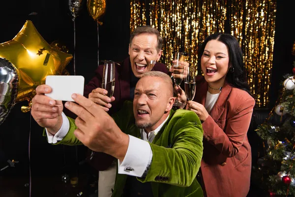 Interracial friends with champagne taking selfie on smartphone near festive decor on black background — Stock Photo