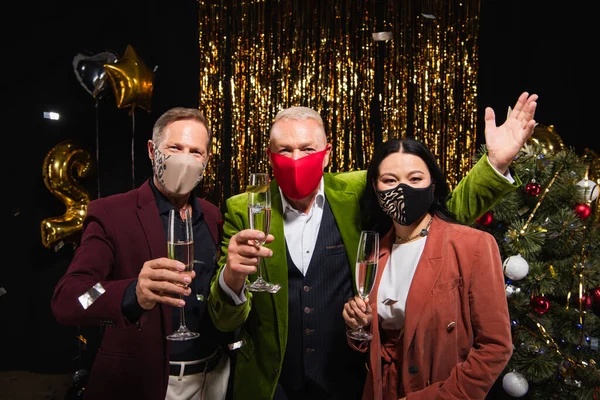 Amigos interraciales en máscaras protectoras sosteniendo copas de champán durante la fiesta de año nuevo sobre fondo negro - foto de stock