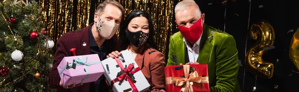 Interracial mature friends in protective masks holing presents near christmas tree on black background, banner — Stock Photo