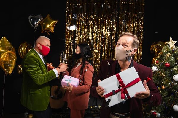 Mature man in protective mask holding present near interracial friends with champagne during new year party on black background — Stock Photo
