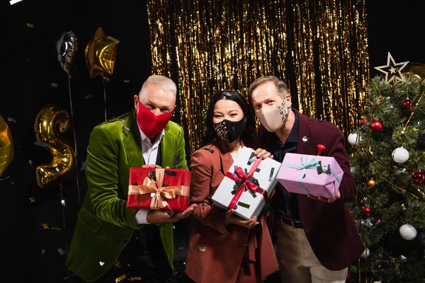 Interracial amigos maduros em máscaras protetoras segurando presentes perto de árvore de natal e balões no fundo preto — Fotografia de Stock