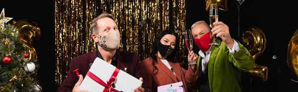 Mature man holding present near interracial friends in protective masks with champagne during new year celebration on black background, banner — Stock Photo