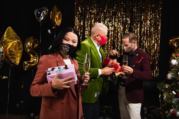 Mature asian woman in protective mask holding champagne and present near friends during new year party on black background — Stock Photo