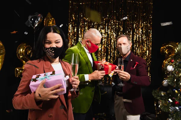 Asian woman in protective mask holding champagne and gift near friends, confetti and christmas tree on black background — Stock Photo