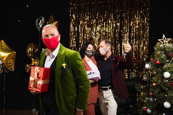 Reifer Mann in Schutzmaske hält Geschenk in der Nähe multiethnischer Freunde mit Champagner und Weihnachtsbaum auf schwarzem Hintergrund — Stockfoto