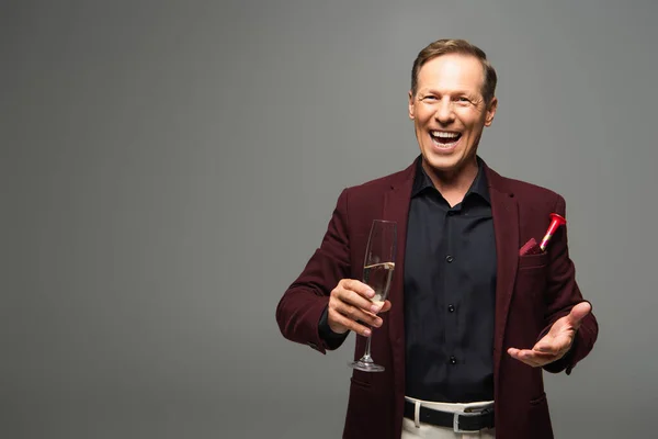 Cheerful man in formal wear holding champagne and pointing at camera isolated on grey — Stock Photo