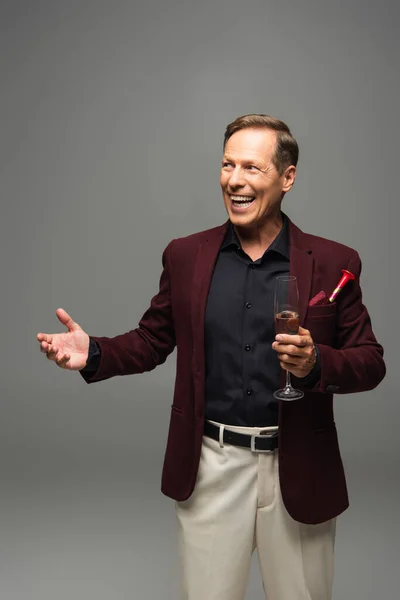 Mature man with champagne and party horn in pocket of jacket pointing with hand isolated on grey — Stock Photo