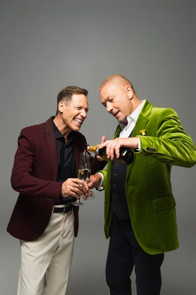 Mature man pouring champagne in glasses near friend in formal wear isolated on grey — Stock Photo