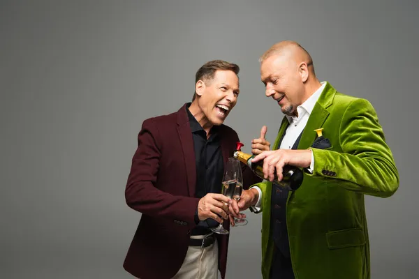 Smiling man pouring champagne in glasses near positive friend showing like isolated on grey — Stock Photo