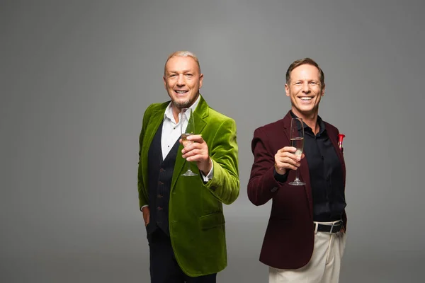 Hombres maduros sonrientes en ropa formal sosteniendo champán aislado en gris - foto de stock