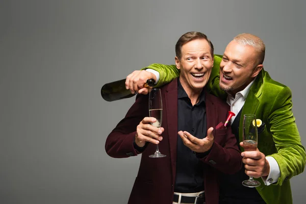 Mature man pointing at glass of champagne near friend with bottle isolated on grey — Stock Photo