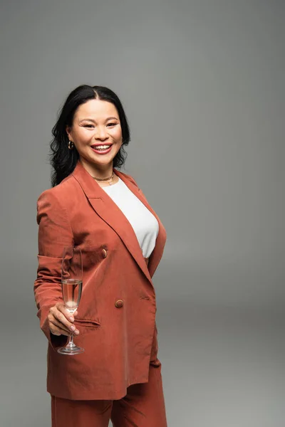 Усміхнена азіатська жінка в куртці тримає келих шампанського ізольовано на сірому — стокове фото