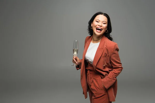 Eccitato asiatico donna in giacca holding champagne e guardando fotocamera isolato su grigio — Foto stock