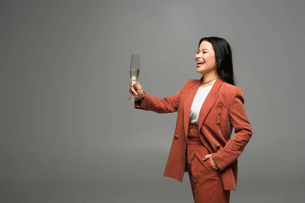 Mujer asiática sonriente en chaqueta de terracota sosteniendo copa de champán aislado en gris - foto de stock