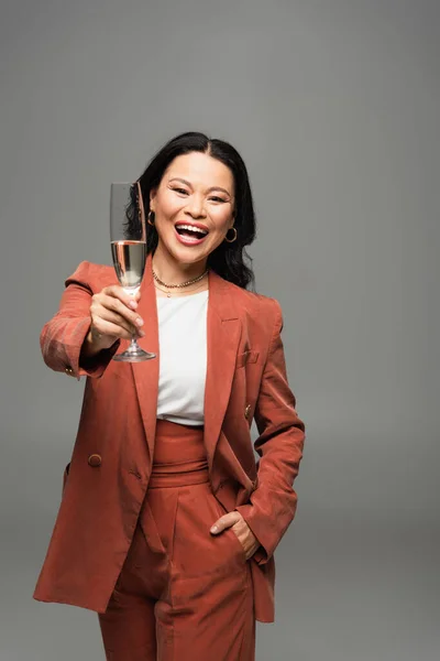 Позитивна азіатська жінка в теракотовій куртці тримає келих шампанського ізольовано на сірому — стокове фото
