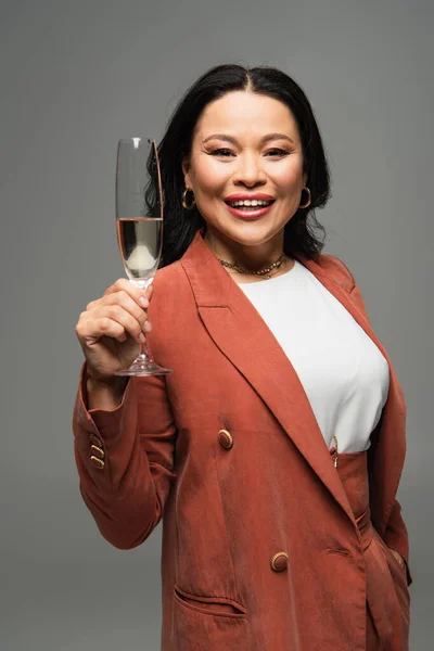Зріла азіатка тримає келих шампанського і посміхається на камеру ізольовано на сірому — стокове фото