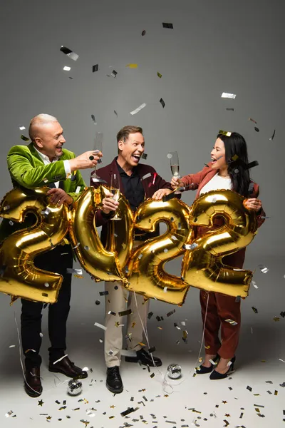 Excited mature friends holding glasses of champagne near balloons in shape of 2022 and confetti on grey background — Stock Photo