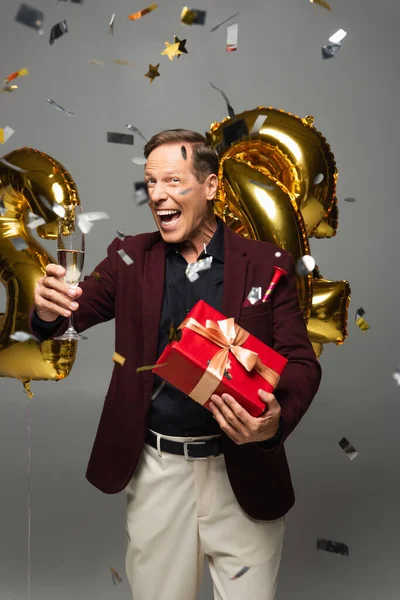 Positive mature man in formal wear holding present and champagne near balloons and confetti on grey background — Stock Photo