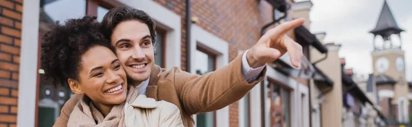 Happy man pointing away with finger near african american girlfriend outdoors, banner — Stock Photo