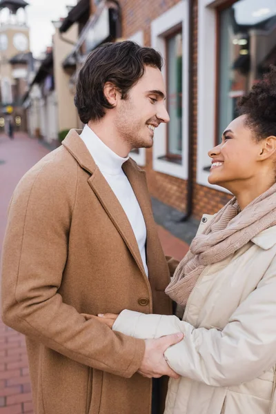 Vue latérale de l'homme heureux souriant tout en embrassant la petite amie afro-américaine à l'extérieur — Photo de stock