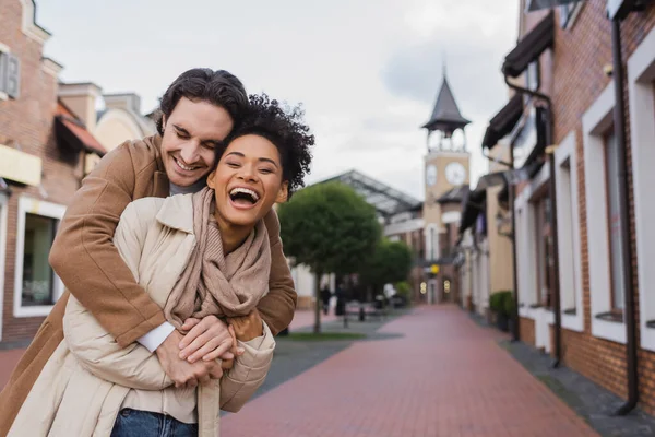 Feliz hombre sonriendo mientras abraza asombrado afroamericano novia al aire libre - foto de stock