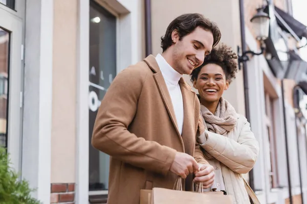 Mujer afroamericana positiva sonriendo cerca de novio sosteniendo bolsas de compras en el centro comercial - foto de stock