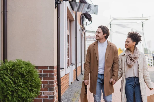 Alegre afroamericana mujer cogida de la mano con novio sosteniendo bolsas de compras mientras camina cerca del centro comercial - foto de stock