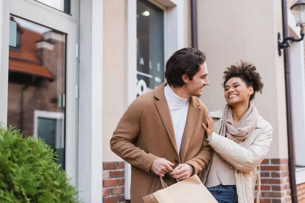 Cheerful african american woman looking at boyfriend with shopping bags near mall — Stock Photo