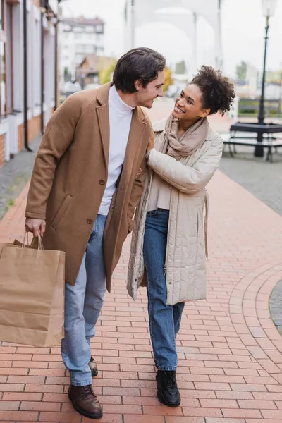 Comprimento total de mulher americana africana alegre olhando para o namorado com sacos de compras perto do shopping — Fotografia de Stock