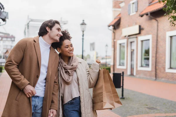 Alegre afroamericana mujer sosteniendo bolsas de compras cerca de novio al aire libre - foto de stock