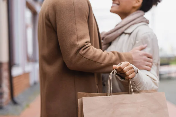 Recortado vista de alegre africana americana mujer sosteniendo bolsas de compras cerca novio al aire libre - foto de stock