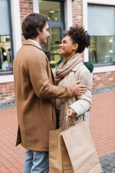 Cheerful african american woman holding shopping bags and looking at boyfriend outdoors — Stock Photo