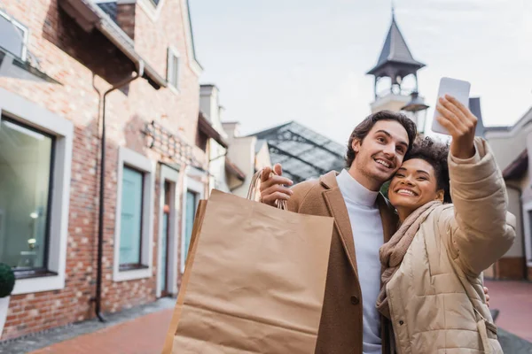 Mujer afroamericana feliz tomando selfie con novio sosteniendo bolsas de compras - foto de stock