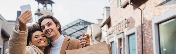 Felice donna afro-americana che si fa selfie con il fidanzato in possesso di borse della spesa, banner — Foto stock