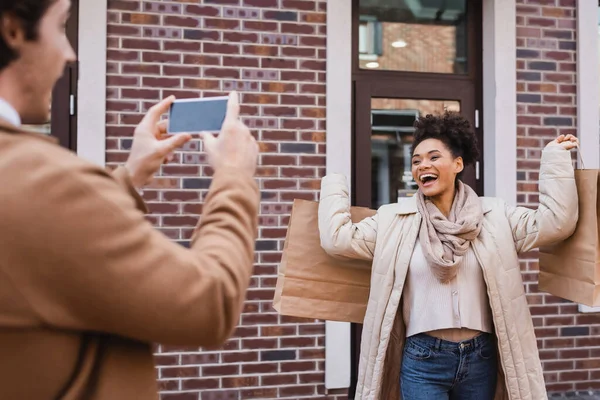 Uomo sfocato scattare foto di donna afro-americana eccitato con borse della spesa — Foto stock