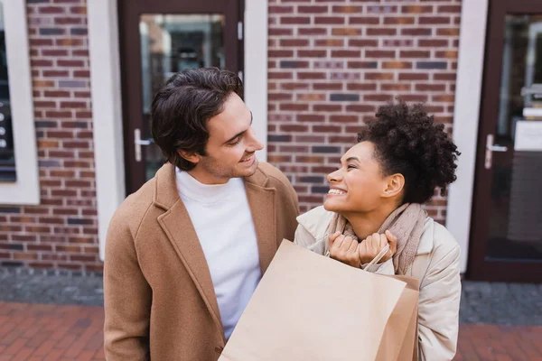 Hombre feliz mirando a la mujer afroamericana complacida con bolsas de compras - foto de stock