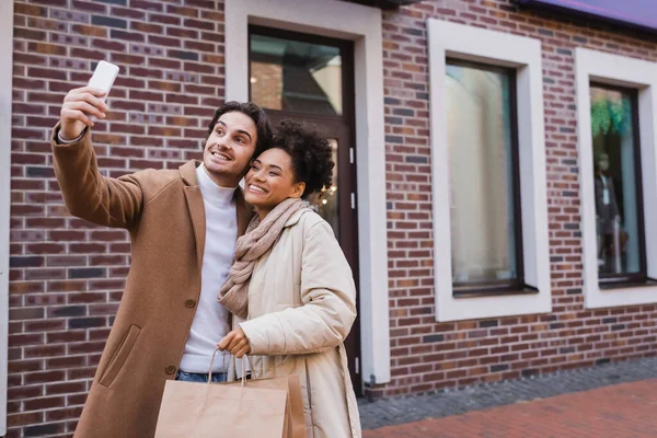 Happy man taking selfie with african american girlfriend holding shopping bags — Stock Photo
