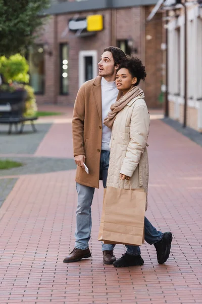 Comprimento total do homem com smartphone em pé perto da namorada americana africana segurando sacos de compras — Fotografia de Stock