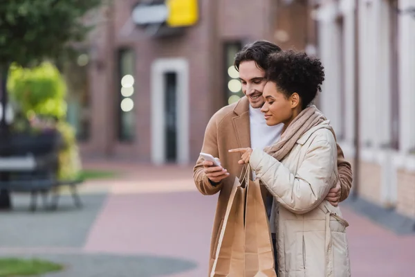 Junge Afroamerikanerin zeigt auf Smartphone in der Nähe ihres Freundes, der Einkaufstüten hält — Stockfoto